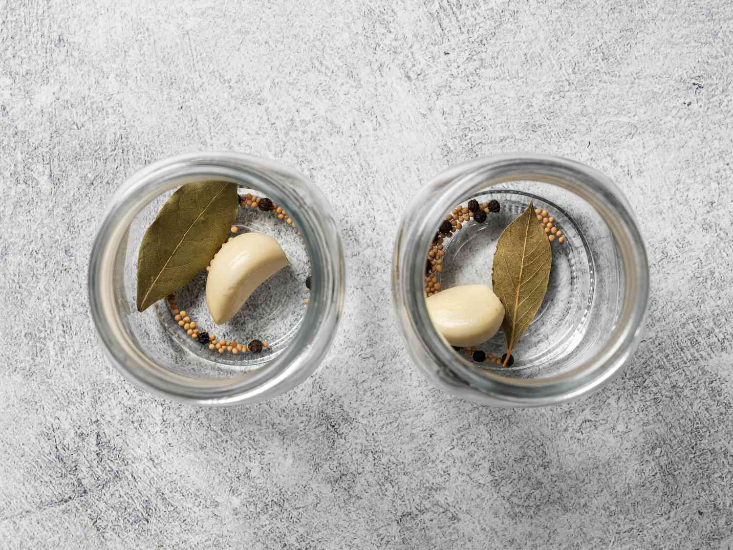 Two glass jars set side by side. Each jar holds a bay leaf, a whole clove of garlic, peppercorns, and mustard seeds.