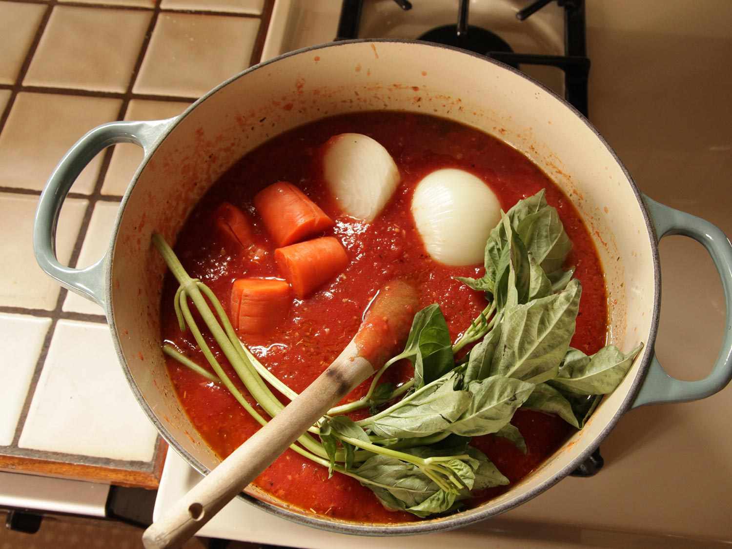 Overhead view of a tomato sauce simmering in a Dutch oven. Several large sprigs of basil have just been added to the pot. 