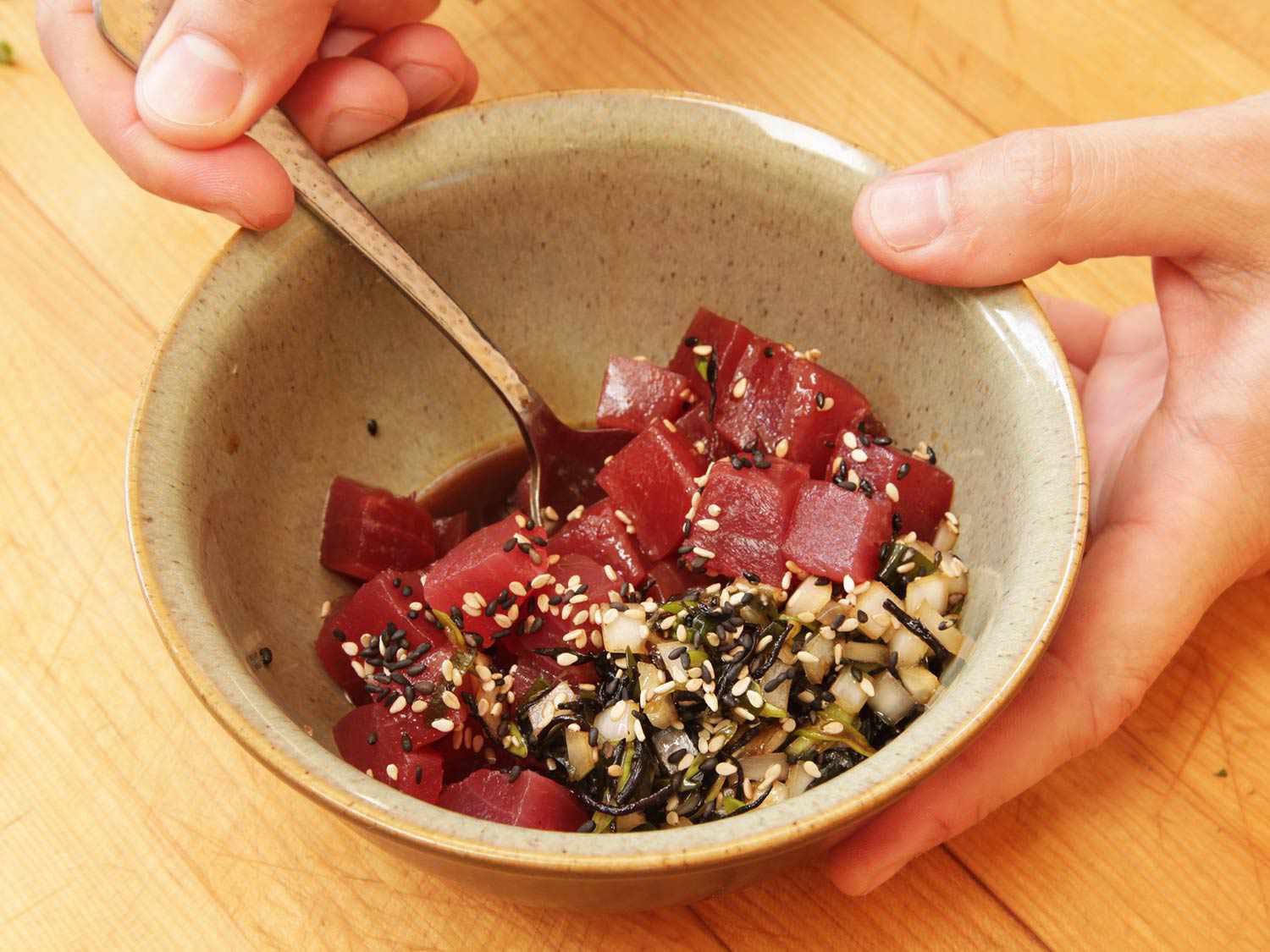 A bowl of cubed tuna and diced onions being dressed with a mixture of soy sauce, sesame oil, and sesame seeds.