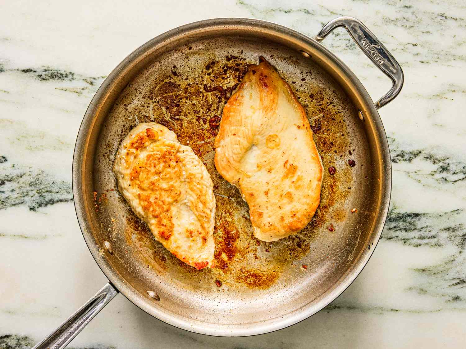 Two pieces of cooked chicken breast in a metal skillet on a marble counter