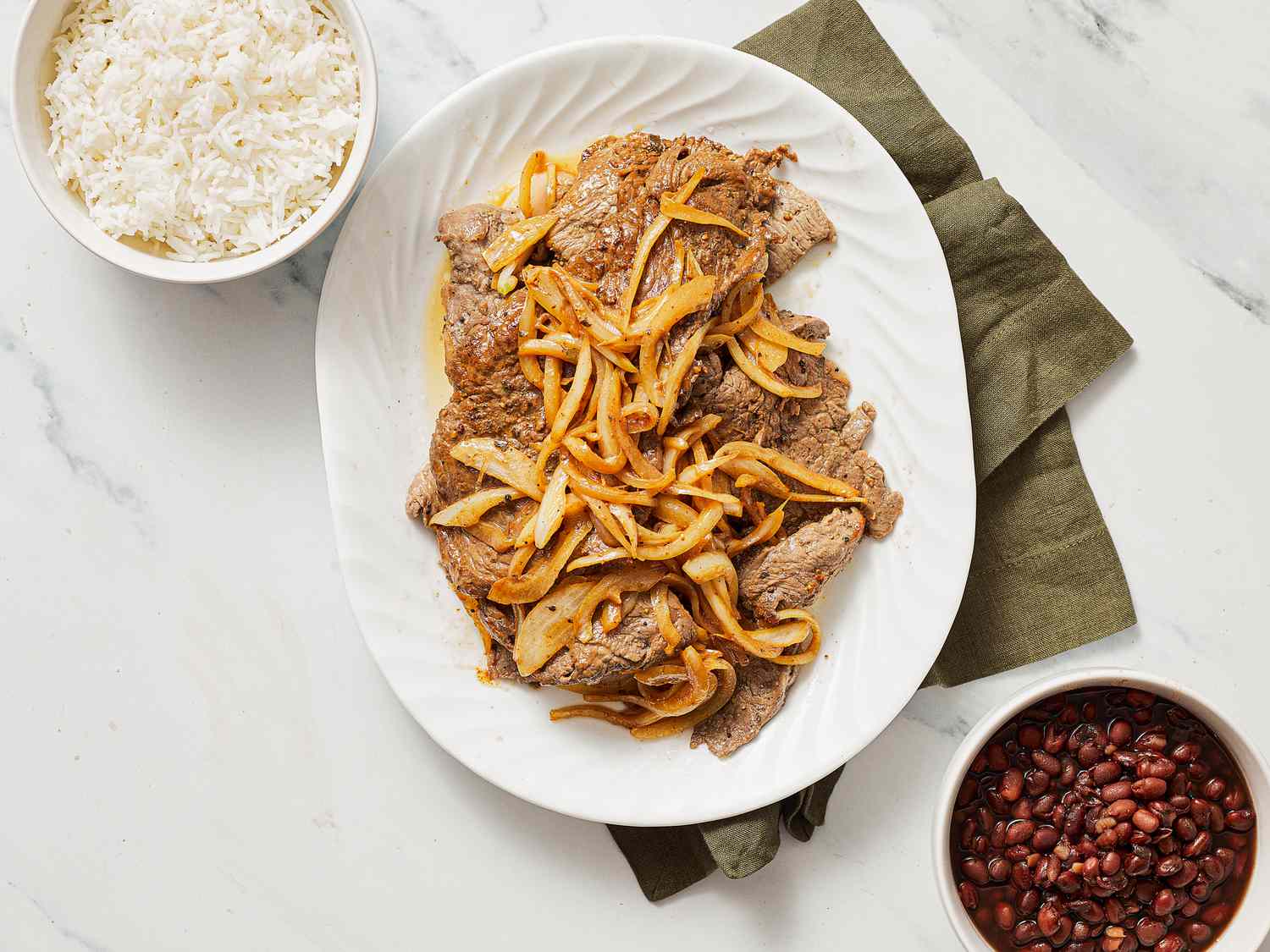 A plate of steak with sautéed onions next to bowls of rice and beans in a kitchen setting