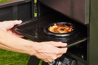 A person adding wood chips to the fuel chamber of the Cuisinart Vertical Propane Smoker