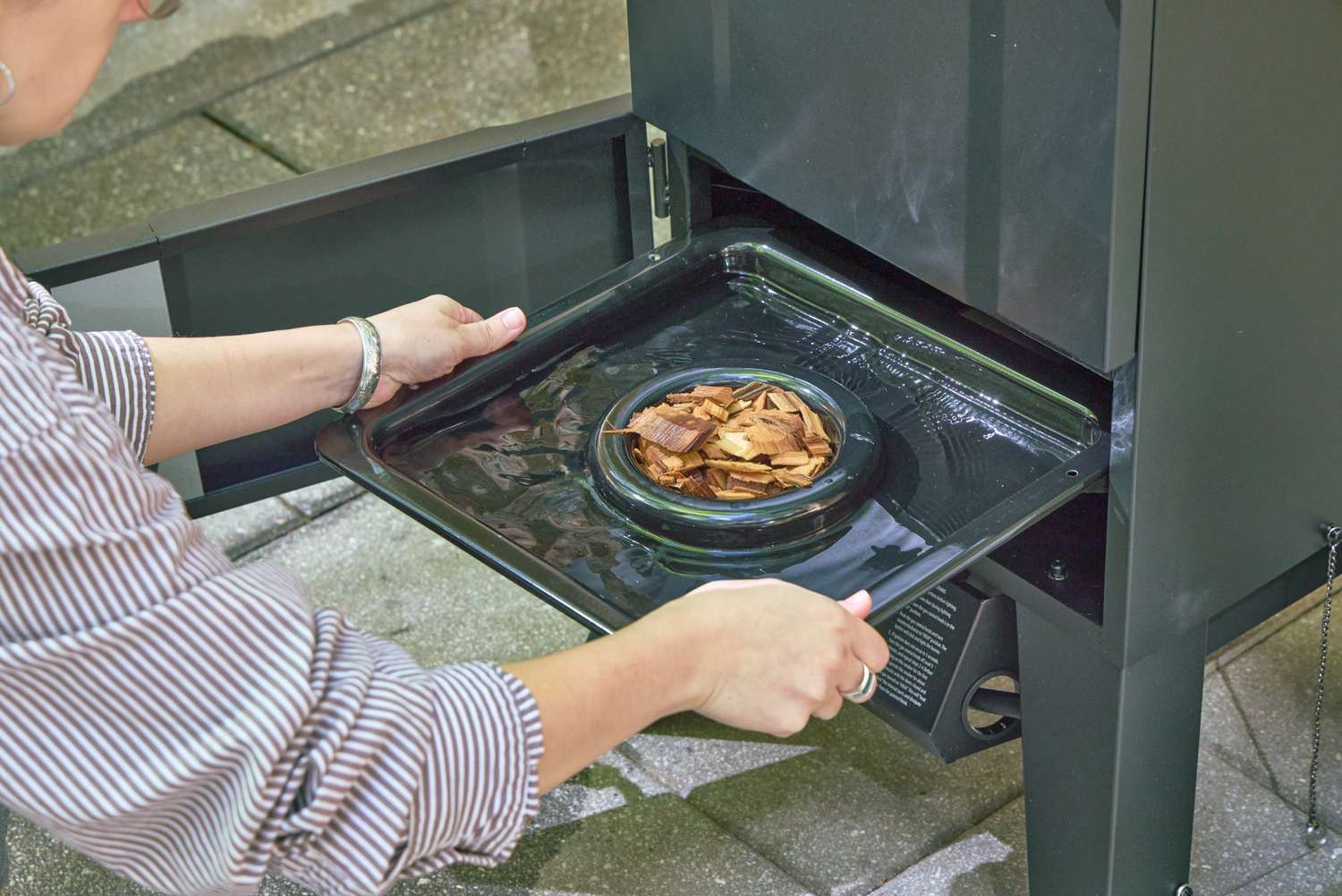 A person places wood chips into the Cuisinart COS-244 Vertical 36-inch Propane Smoker