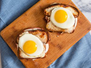 Two Croque Madame sandwiches, topped with sunny-side up eggs on a wooden board on a blue cloth.