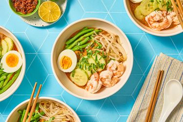 Bowls of curry laksa garnished with shrimp boiled egg cucumber and beans accompanied by lime and chili paste on a blue background
