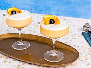 Two whiskey sour cocktails, garnished with an orange wheel and a cherry on a cocktail pick, served on a tray