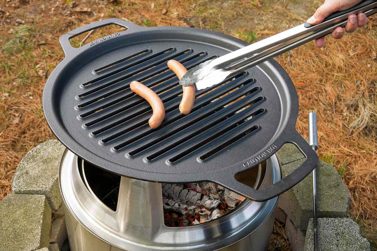 Cooking hot dogs on a grill over a fire pit using tongs