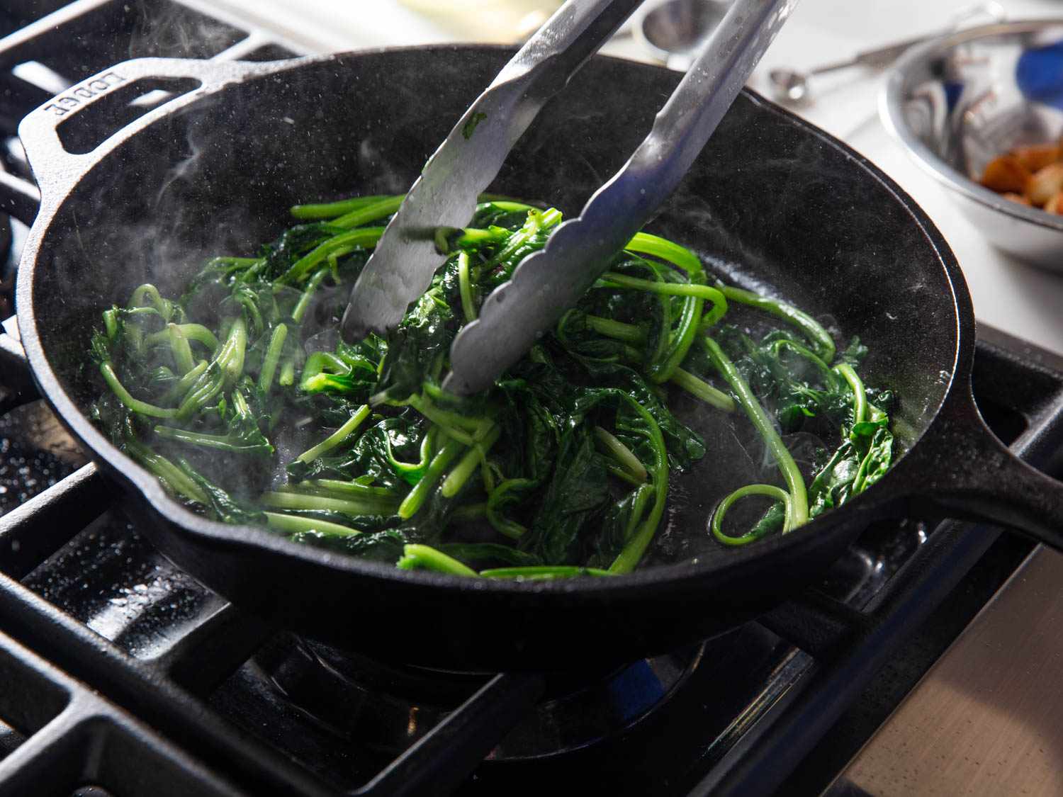 Spinach is sautéed in the skillet and stirred with a pair of tongs.