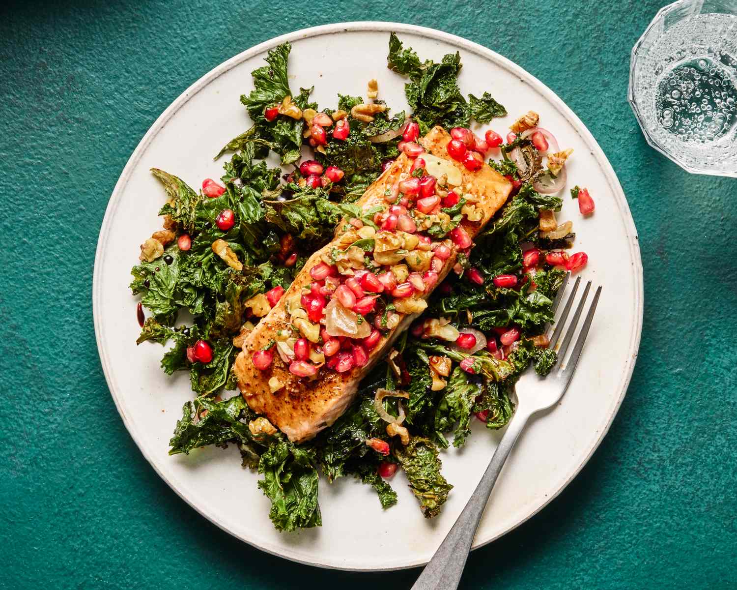 A dish of salmon glazed with pomegranate sauce served on a bed of kale and garnished with pomegranate seeds and nuts with a fork