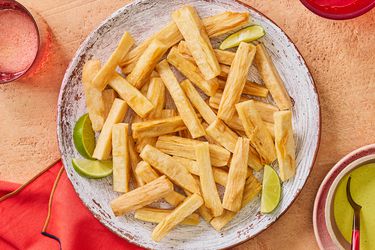 Fried yuca on a rustic plate, and textured terra cotta surface, with limes, dipping sauce and beverage on the side 