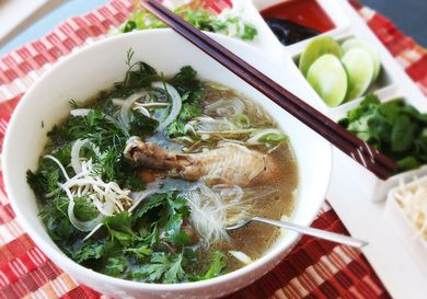 White bowl of pho ga on red placemat with chopsticks and a spoon on the bowl and garnishes on the side.