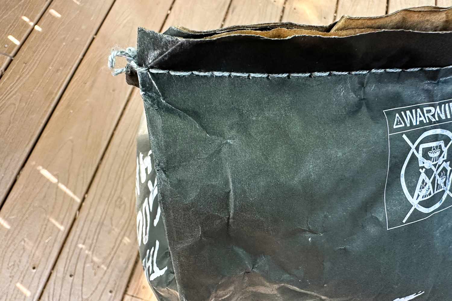 A partially open black paper bag of The Good Charcoal Company Premium Hardwood Lump Charcoal with warning symbols on the side placed on a wooden surface