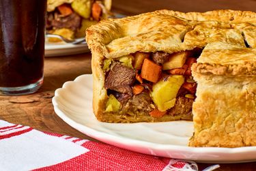 A meat and vegetable potpie with a sliced section showing the filling served on a white plate with a drink in the background
