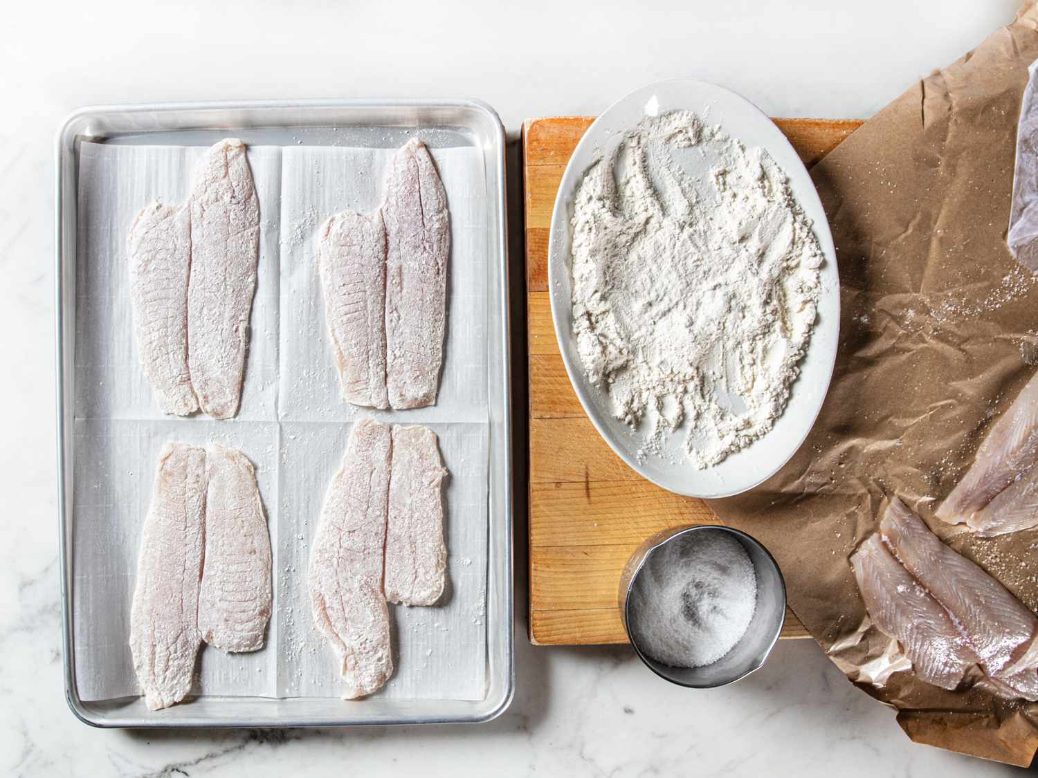 Four pieces of sole, dredged in flour on a baking sheet lined with parchment paper. Next to the baking sheet on a wooden cutting board is the plate with flour a bowl of salt and the remaining pieces of sole.