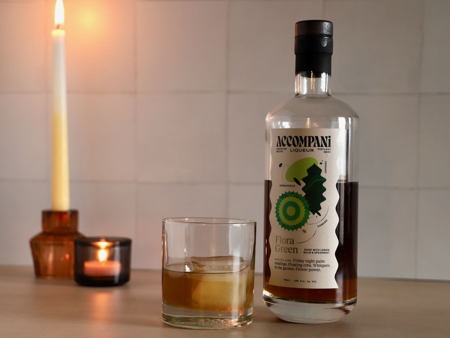 A bottle of Accompani Flora Green Liqueur beside a glass containing an iced beverage with lit candles in the background