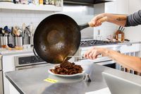 Person pouring food from a Mammafong Pre-Seasoned Blue Carbon Steel Flat-Bottom Pow Wok onto a plate
