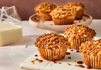 apple pie muffins on a white tray, with 3 muffins on a circular tray in the background. A clear glass of milk to the side on pink napkins. 