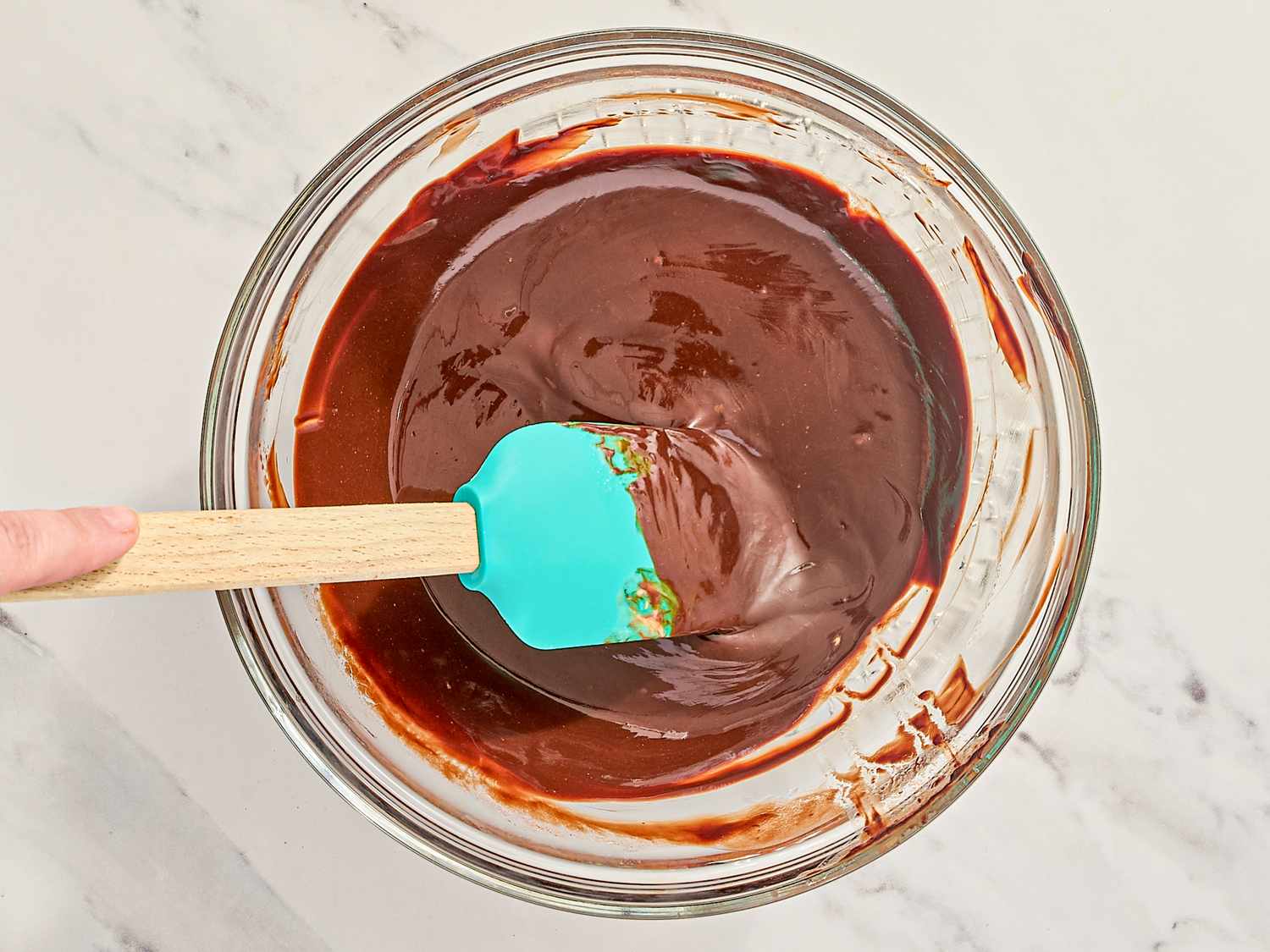 A glass bowl containing chocolate mixture being stirred with a blue spatula