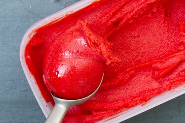 A rectangular container of bright red strawberry sorbet with a scoop being pulled from it.