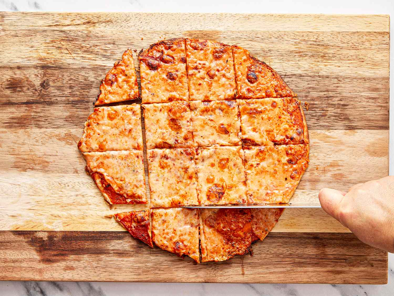 A hand cutting a squaresliced pizza on a wooden board