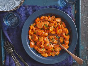 Glazed carrots in a blue bowl on blue rustic surface. A spoon is in the bowl, with a few carrot slices in it, and there is cups of sparkling water, and plates on the side.