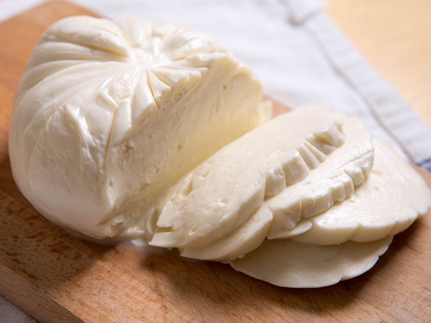 Close-up of a ball of fresh mozzarella on a cutting board. Several slabs of mozzarella have been sliced from the ball and are shingled to one side.