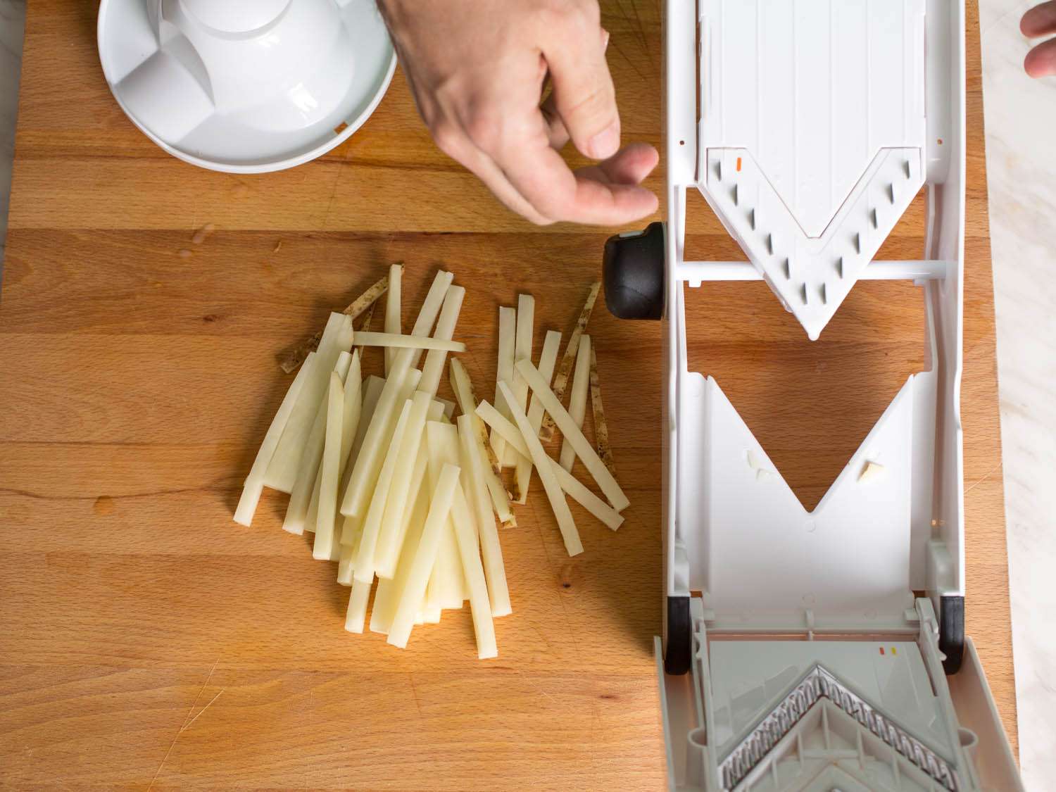 Thin slices of potato next to the OXO Mandoline that is on a wood surface