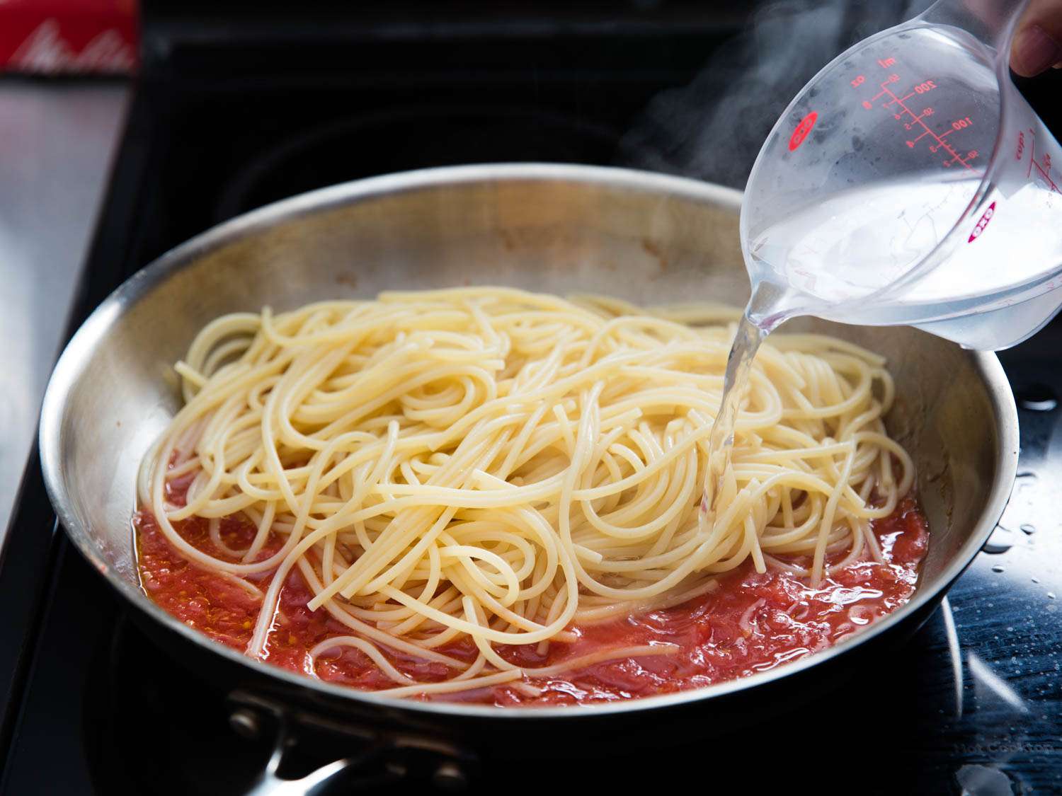 Water from cooking pasta being added to spaghetti and red pasta sauce cooking in a skillet.