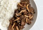 A closeup shot of stir-fried beef with mushrooms and butter. The beef and mushrooms are plated in a round white bowl with white rice on a white tile background, and the shot shows how the mushrooms have been seared, while the feed is tender and in a somewhat glossy sauce.