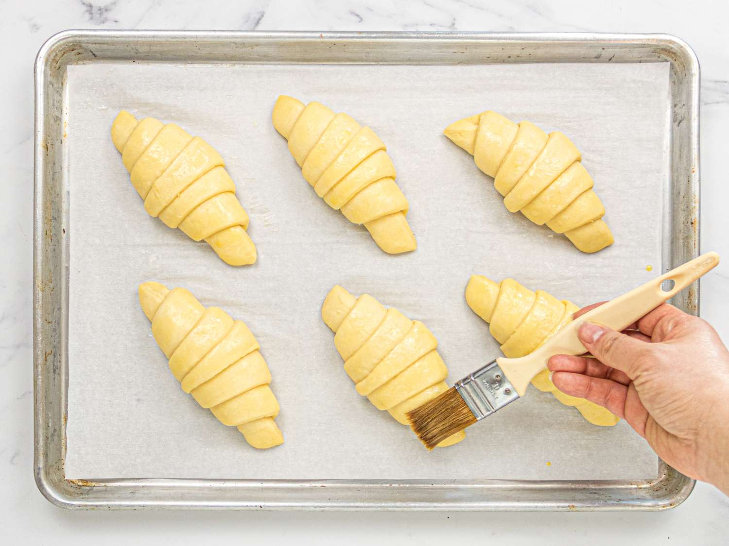 Hand applying egg wash to dough croissants on a baking tray lined with parchment paper