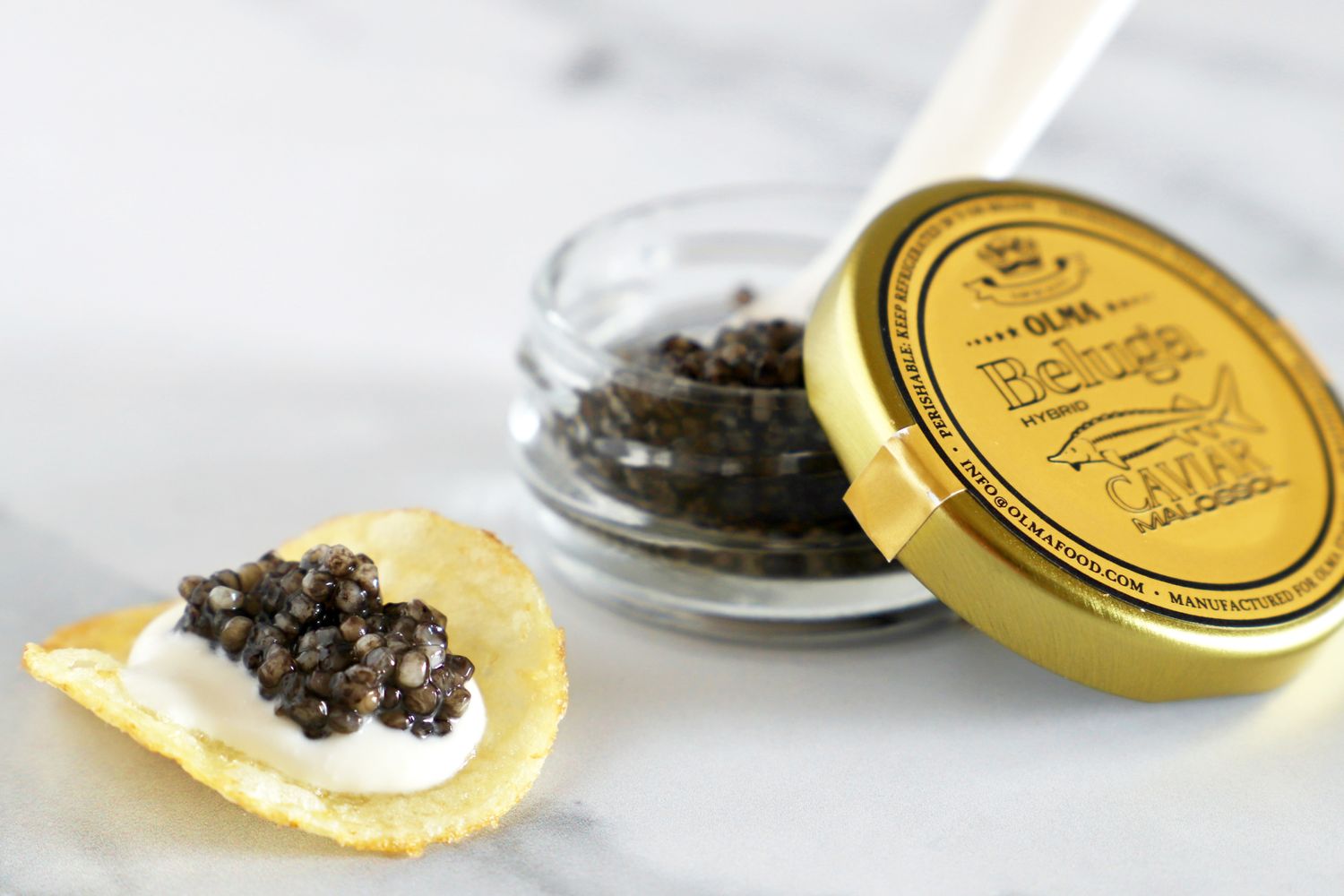 An open jar of beluga hybrid caviar next to a potato chip topped with crème fraîche and a scoop of caviar