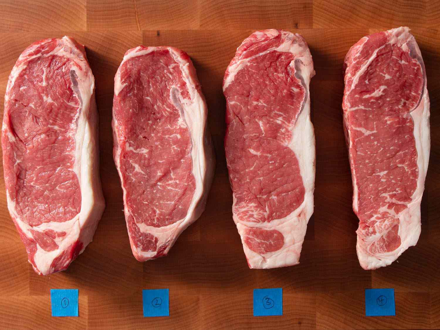 Overhead view of four strip steaks identified by numbered tape on a cutting board