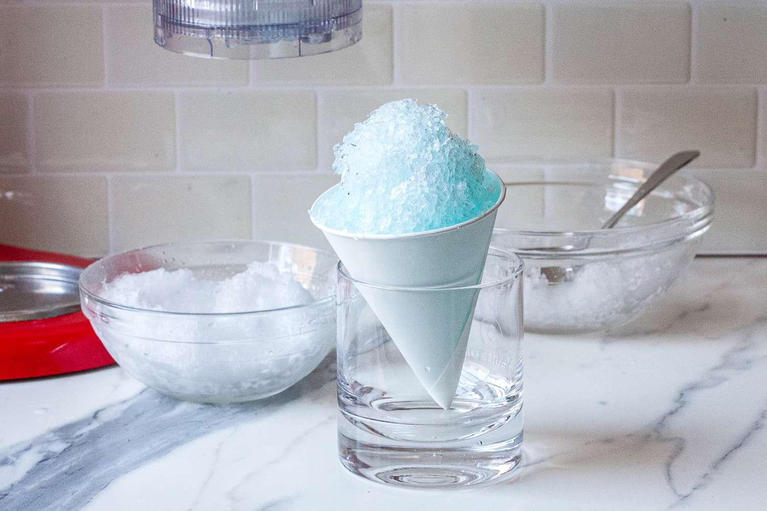 A snow cone with blue syrup beside a bowl with more shaved ice in it.
