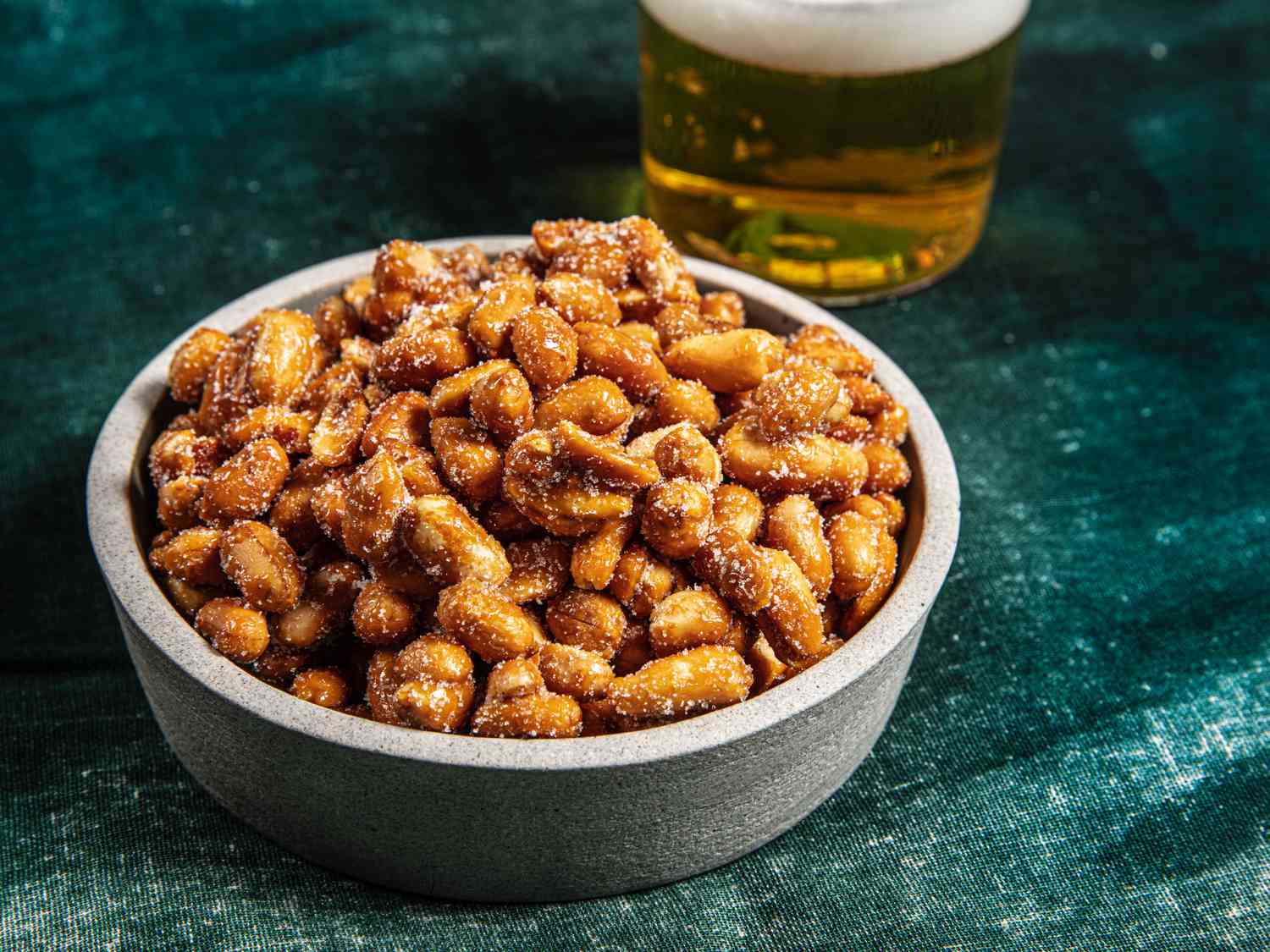 Bowl of honey roasted peanuts on a textured green fabric next to a glass of beer