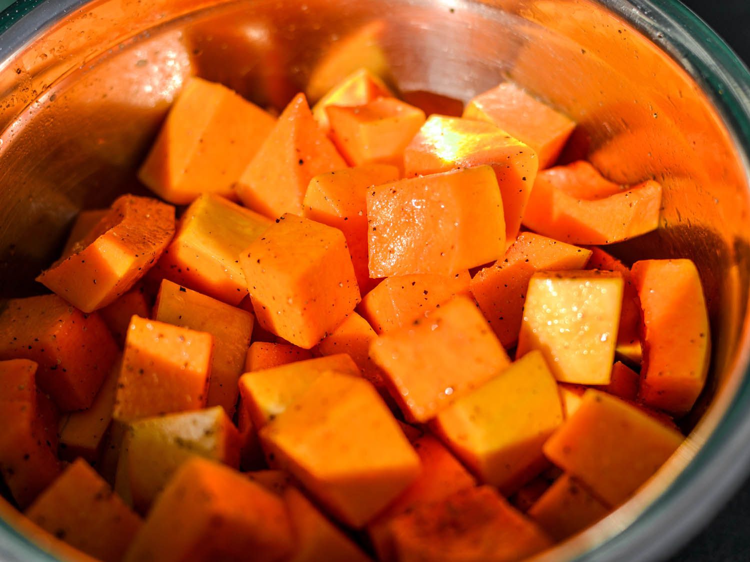 Butternut squash cubes tossed in salt, pepper, and olive oil in a mixing bowl.