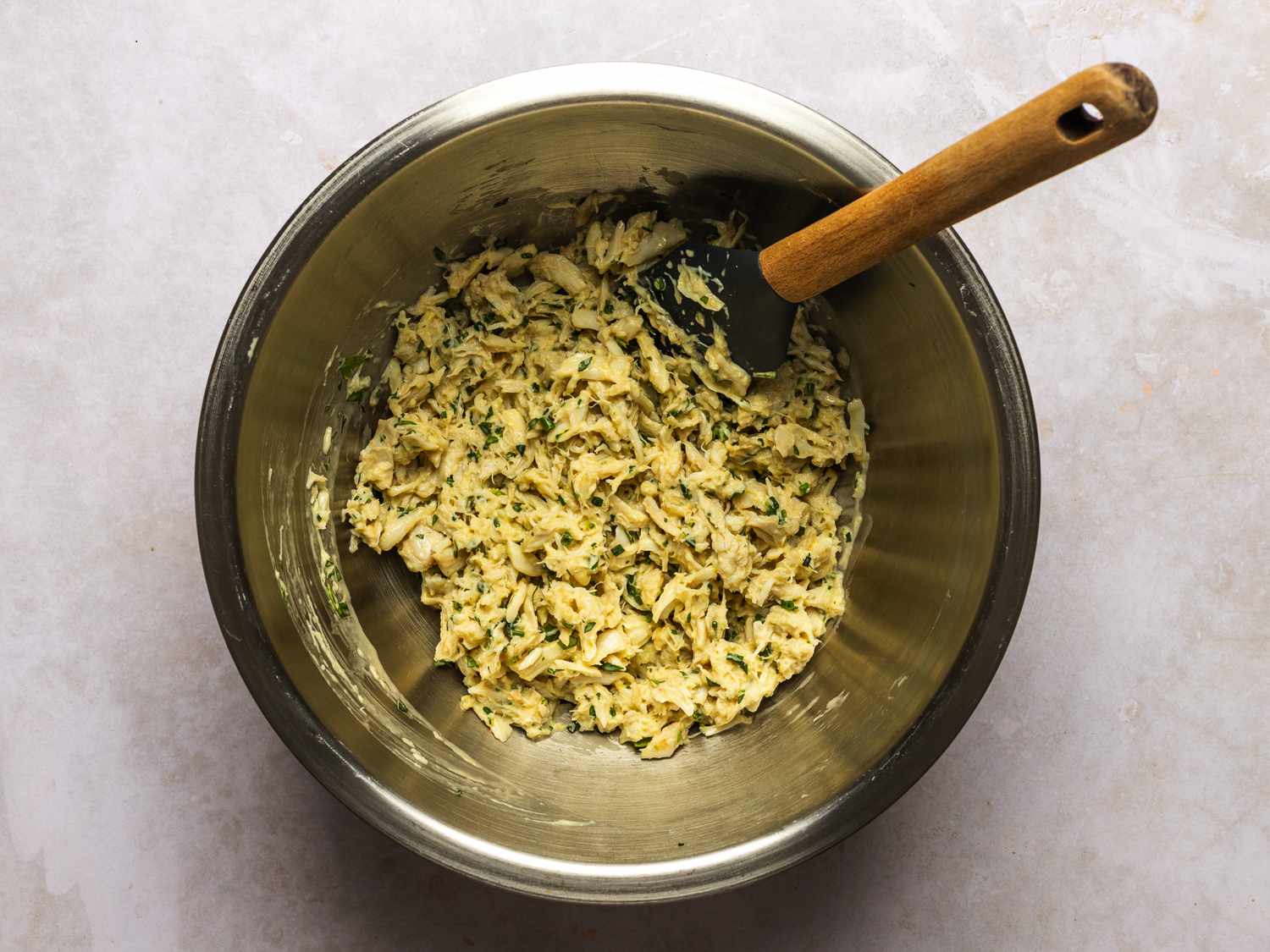Crab, mayonnaise, herbs, and an egg combined inside a large bowl with a flexible spatula.