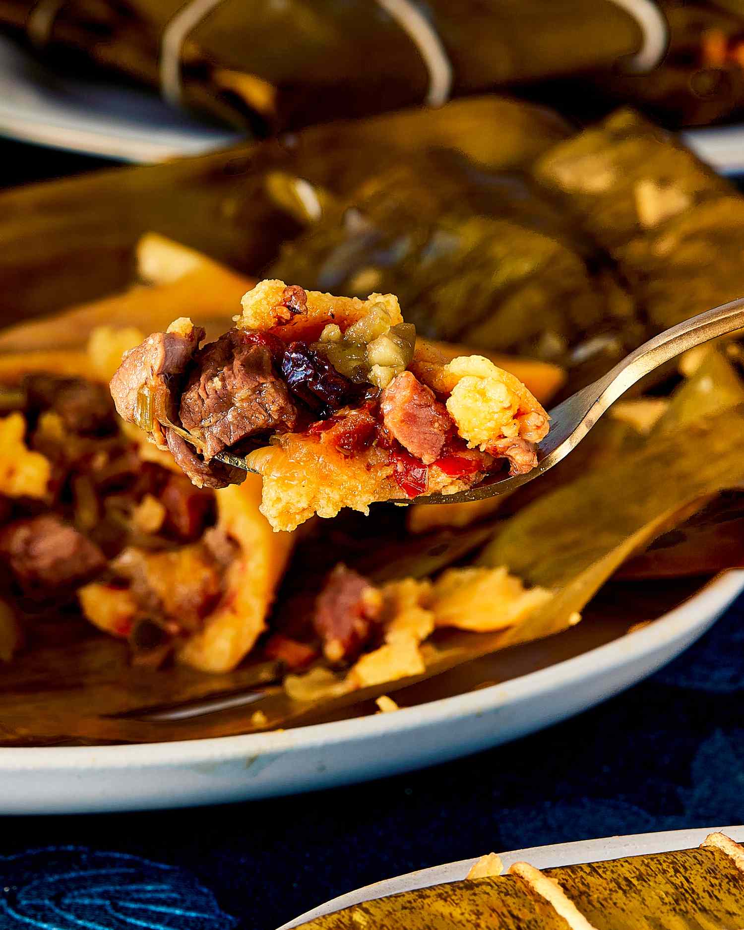 Fork holding a portion of hallacas over a plate showing a mix of meat and ingredients wrapped in a banana leaf