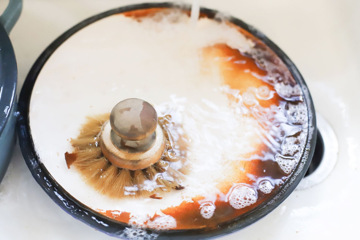 a braiser's lid in the sink being scrubbed by a stiff-bristled brush