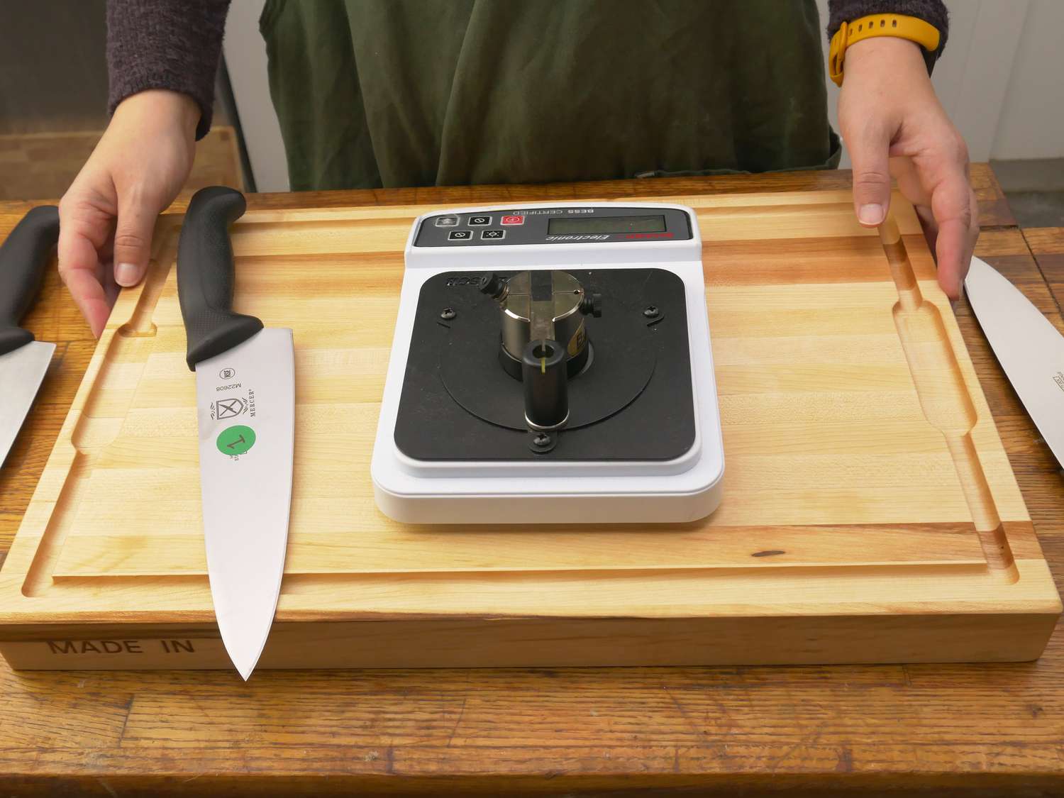 a person with the made in cutting board, a chef knife, and a knife sharpness reader