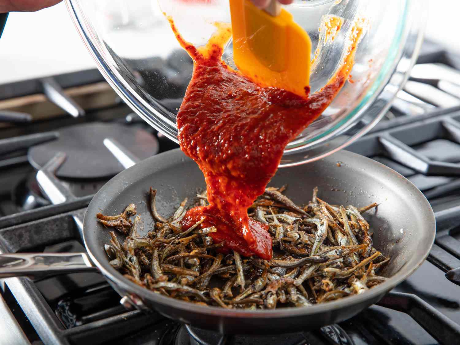 The gochujang mixture is scraped from the mixing bowl with a flexible spatula and added to the pan.