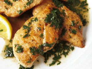 Closeup of chicken cutlets in a pan sauce with fresh herbs and a lemon wedge.