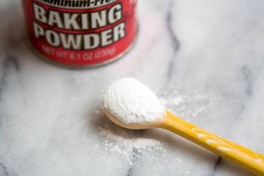 A container of baking powder next to a spoon full of baking powder. 