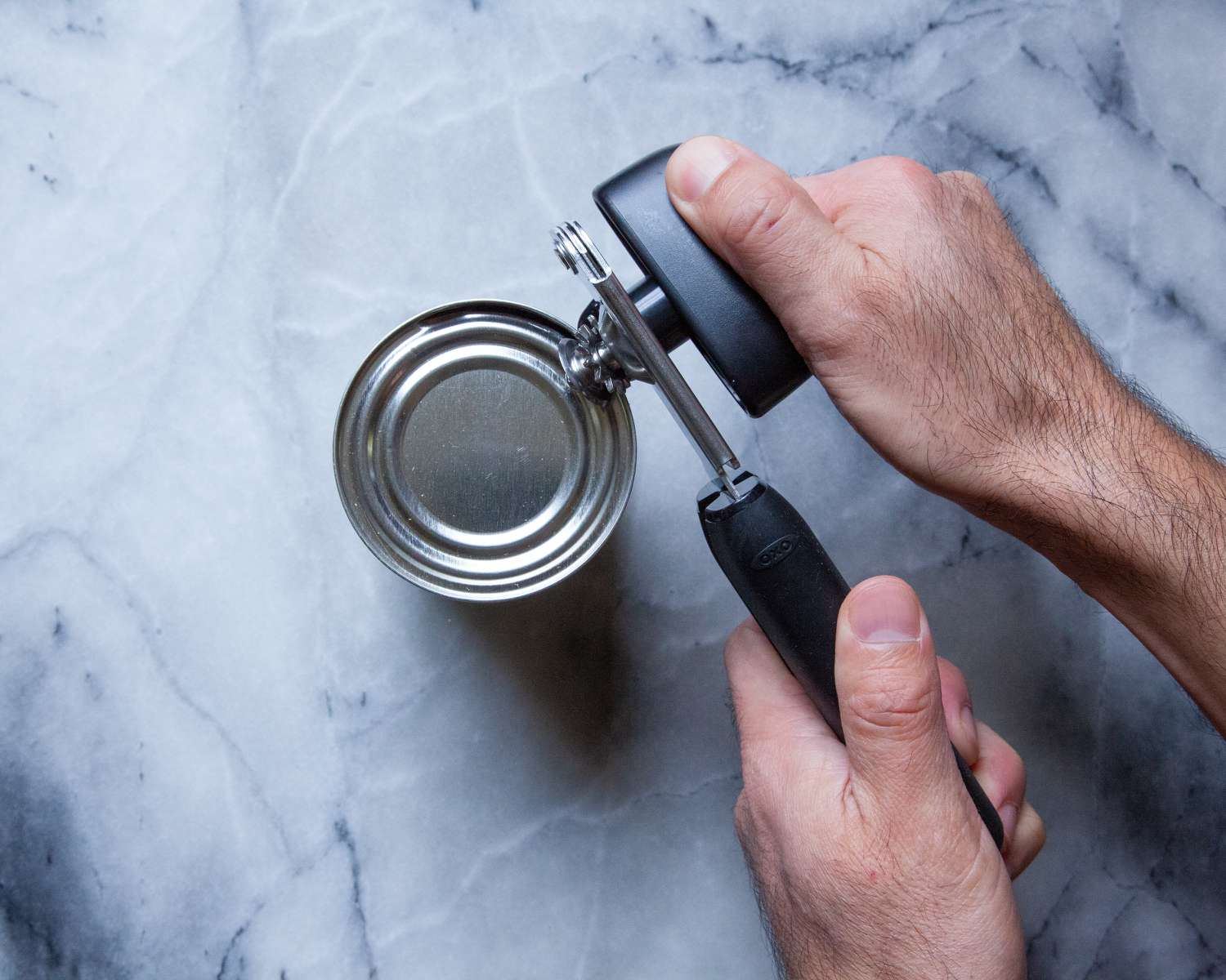A person using the OXO can opener to open a can on a marble countertop.