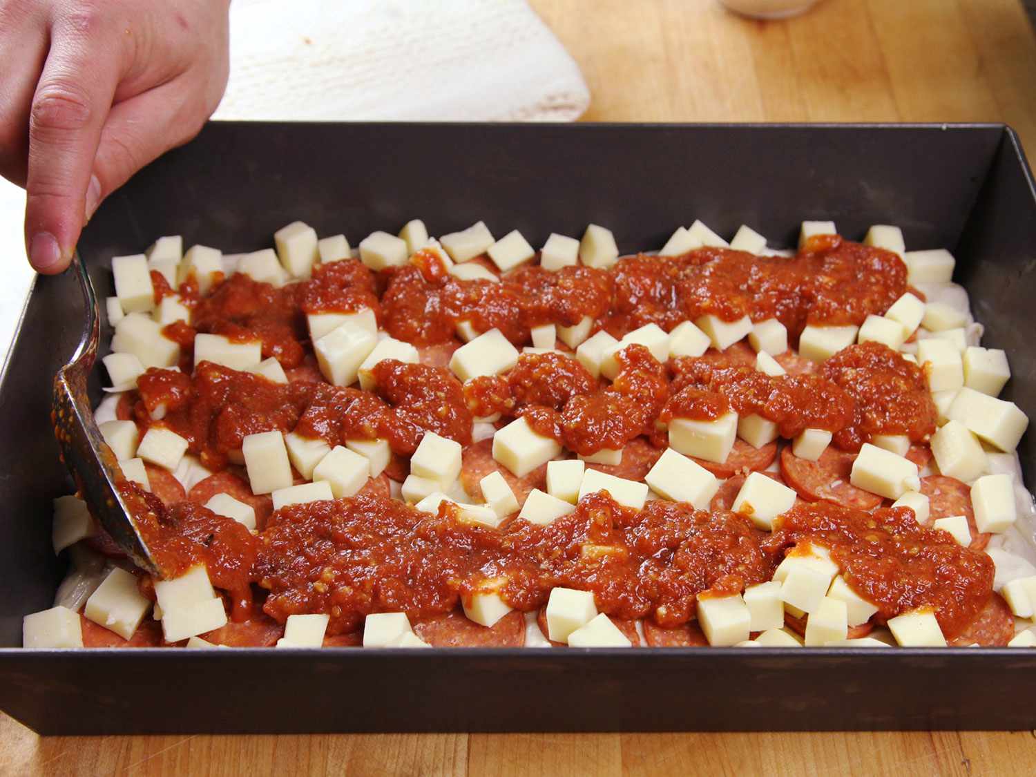 A hand spooning tomato sauce on top of an uncooked Detroit-style pizza. The sauce is applied in three even rows on the top of the pizza. 