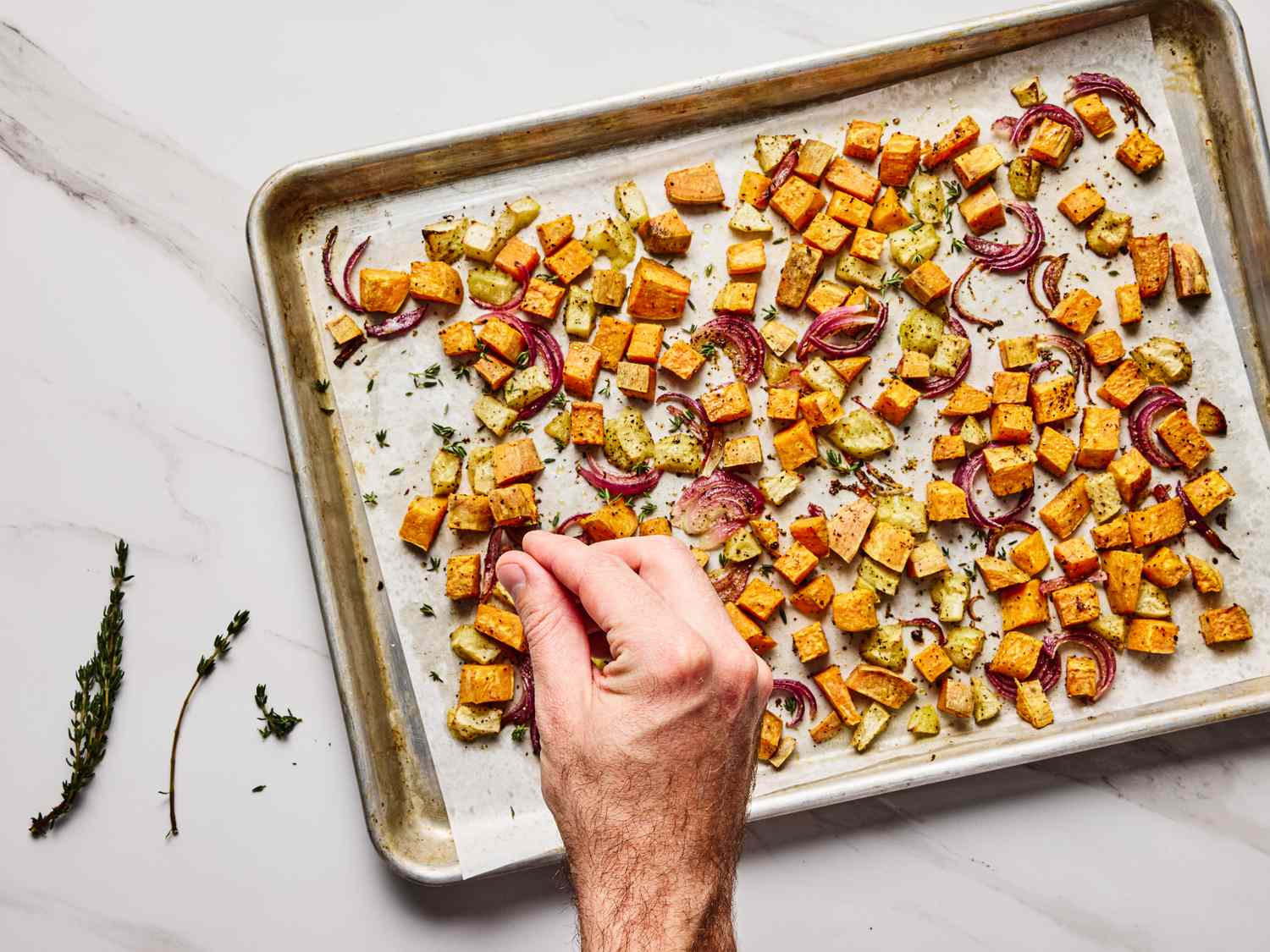 A tray of roasted vegetables including diced sweet potatoes and onions with a hand garnishing them with fresh herbs