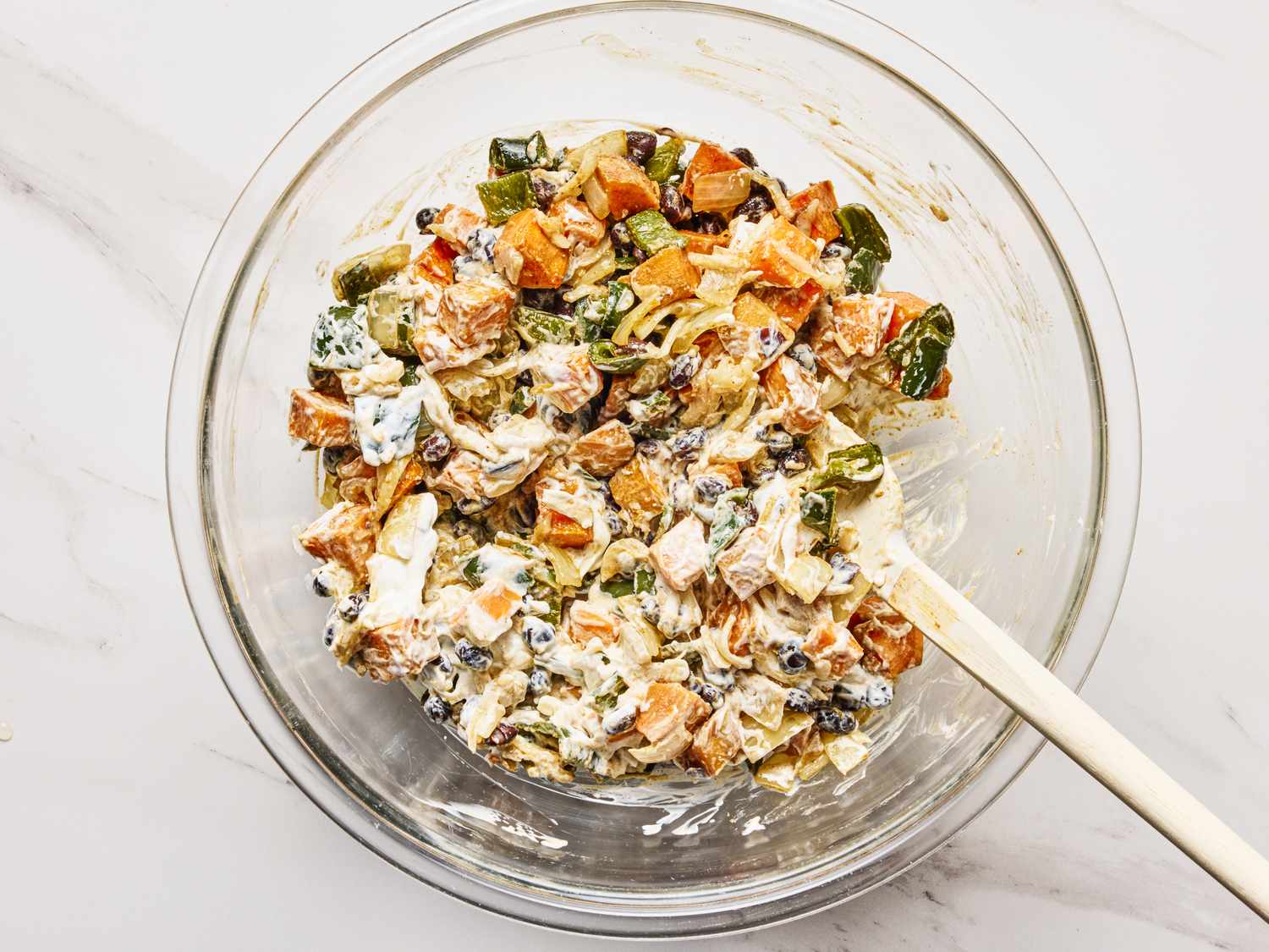 A glass bowl containing a mixed filling for sweet potato enchiladas with diced vegetables and a creamy sauce being stirred with a spatula