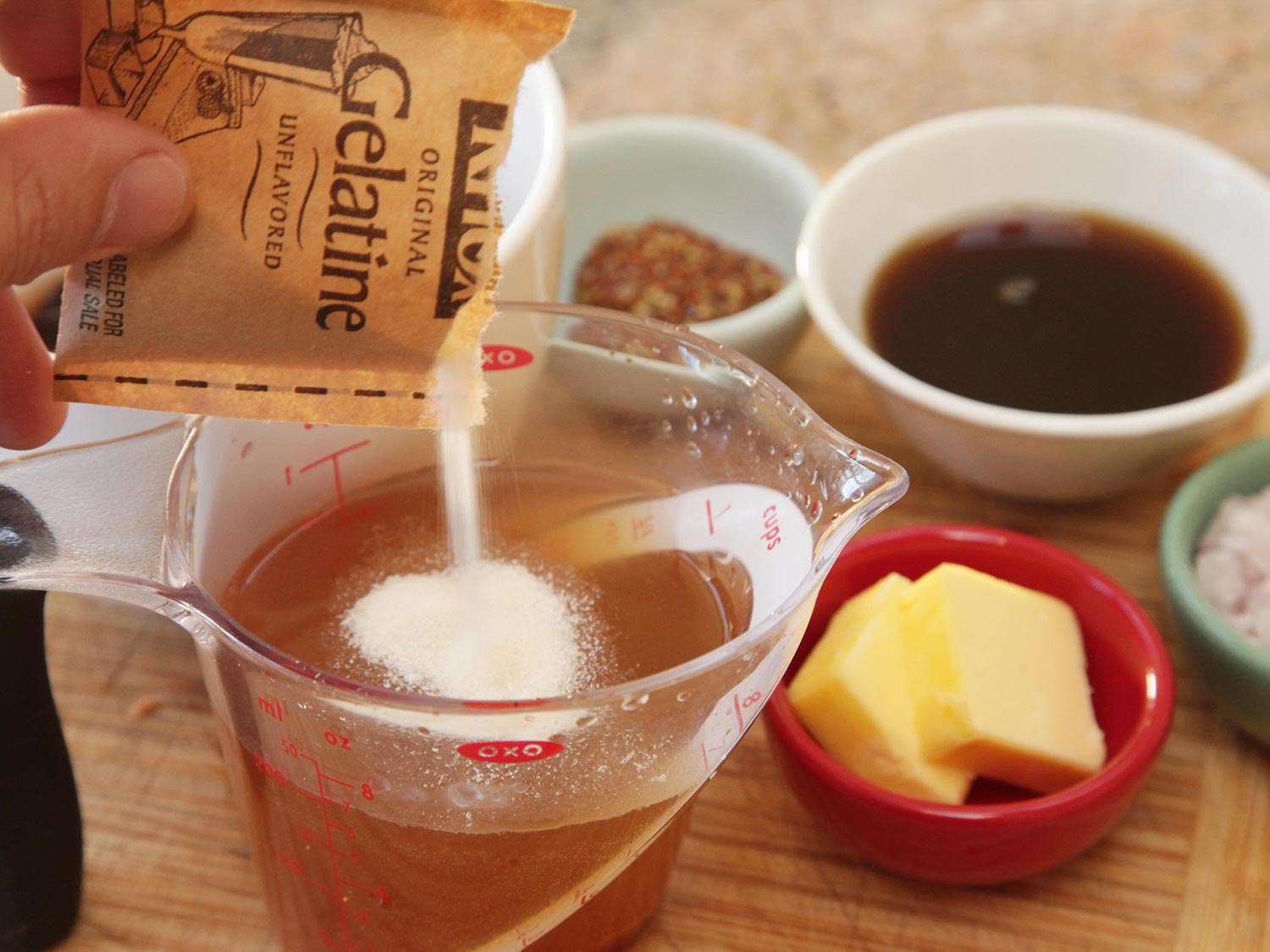 Adding gelatin to a measuring cup containing chicken broth to add body, next to other ingredients for the bourbon glaze.