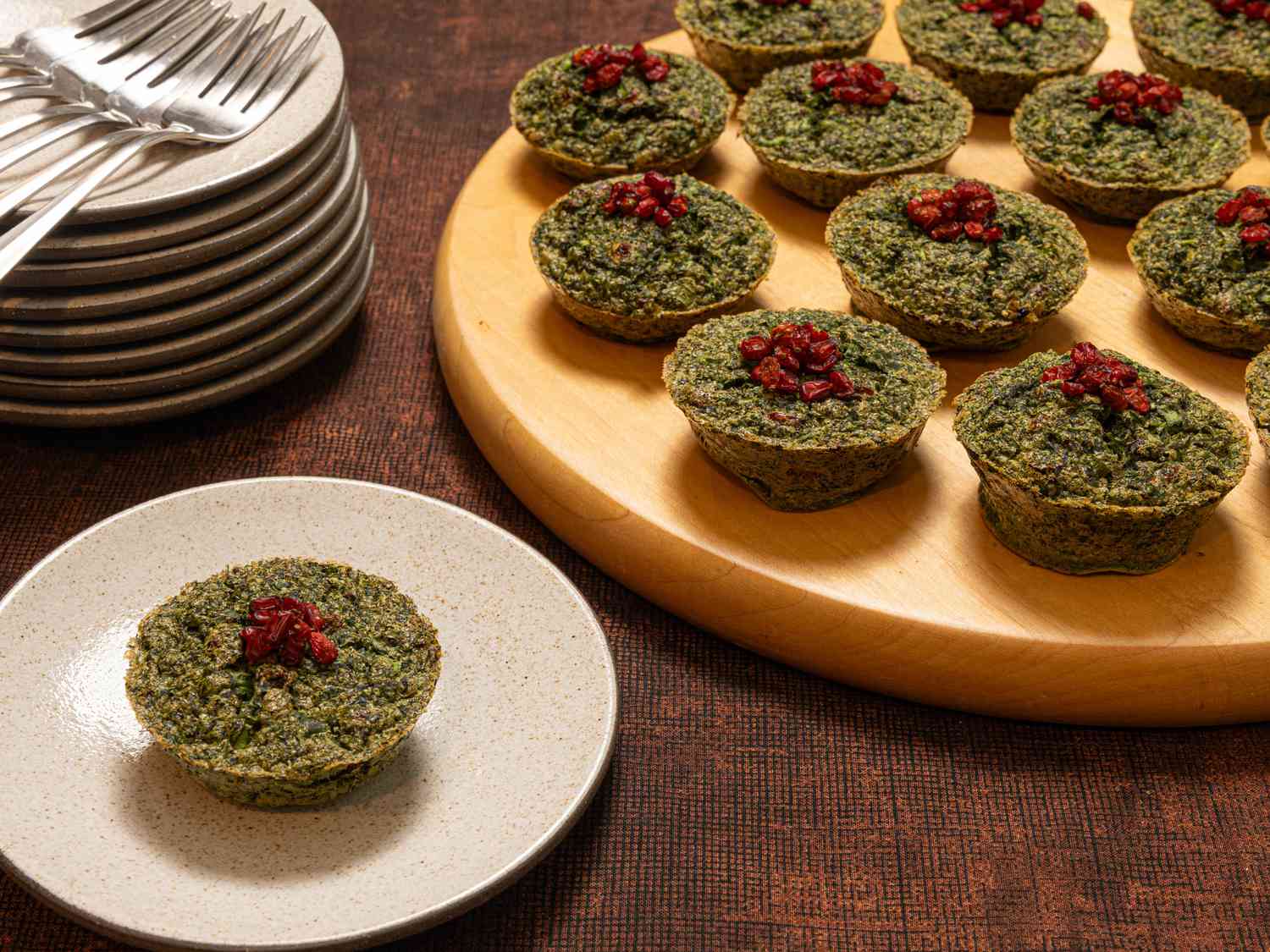 Small circular servings of Herb kookoo on a wooden platter, with a single serving on a small dish. Stack of plates and forks to the side. 