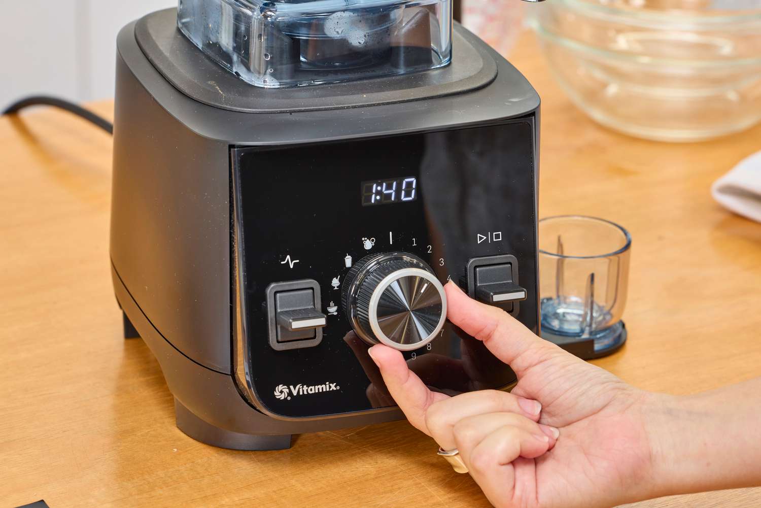 A close-up of a hand turning a nob on the Vitamix Ascent X2 resting on a wooden surface