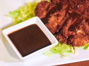 A small white square ramekin of tonkatsu sauce next to a fried cutlet. 